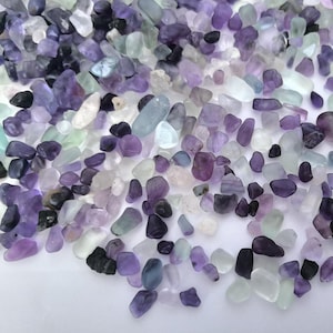 May include: A collection of tumbled fluorite stones in various shades of purple, lavender, white, and green. The stones are smooth and irregular in shape, with a mix of opaque and translucent qualities. The arrangement creates a visually appealing display of natural gemstones.