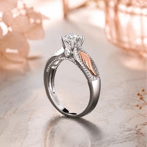 May include: A silver engagement ring with a large, clear gemstone at the center. The ring features a decorative design with a rose gold accent on the side. The band is detailed with small, beaded accents.