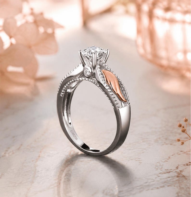 May include: A silver engagement ring with a large, clear gemstone at the center. The ring features a decorative design with a rose gold accent on the side. The band is detailed with small, beaded accents.