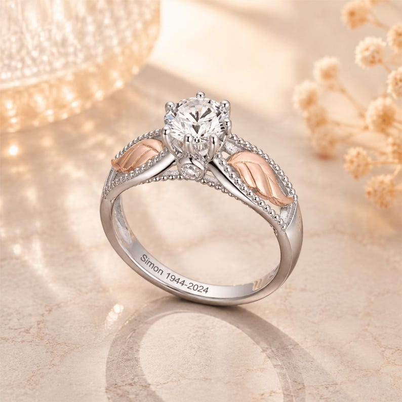 May include: A silver engagement ring with a large, clear gemstone at the center. The ring features rose gold leaf accents on either side of the gemstone and small, decorative beads along the band. The inscription "Simon 1944-2024" is engraved on the band.