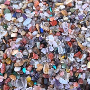 May include: A close-up view of a collection of polished gemstones in various colors, including red, blue, purple, and clear. The gemstones are of different shapes and sizes, creating a textured surface. The image is well-lit, highlighting the natural beauty of the stones.