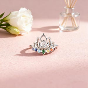 May include: A silver ring with a lotus flower design. The ring features a large, clear, pear-shaped stone at the center and smaller, colorful gemstones in a rainbow arrangement. The ring is set against a soft pink background.