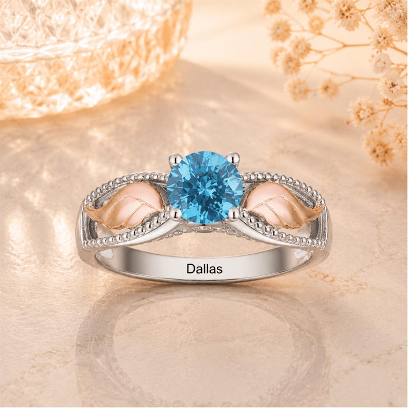 May include: A silver ring with a large, round, light blue gemstone at the center. The ring features two rose gold-colored leaf accents and the name "Dallas" engraved on the band.