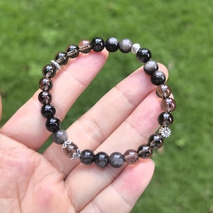 May include: A beaded bracelet featuring dark, smoky quartz beads in shades of black, gray, and brown. The bracelet includes small silver-colored decorative accents, including a flower-shaped charm. The bracelet is held in a hand against a blurred green background.