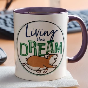 May include: A white ceramic mug with a purple interior and handle. The mug features the text "Living the Dream" in blue and green, along with a cartoon hamster. The mug is on a wooden surface.