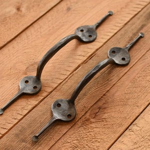May include: Two dark grey metal handles with a hammered texture. Each handle has a decorative, key-shaped end with two holes and a curved bar. The handles are displayed on a wooden surface.