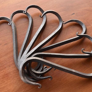 May include: A collection of handcrafted metal hooks, each with a heart-shaped design. The hooks are made of a dark, textured metal and are arranged in a fan-like pattern on a wooden surface. The hooks are approximately 15 cm long.