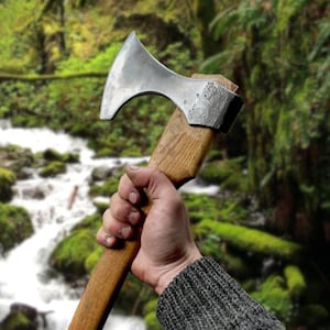 May include: A steel axe with a wooden handle is held in a hand. The axe head is silver and the handle is a light brown color. The background is a blurred image of a forest with a stream.