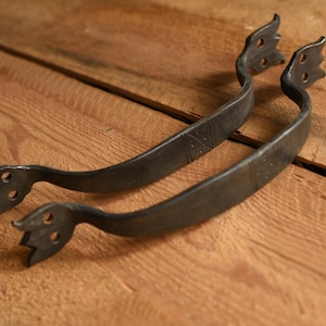 May include: Two rustic, dark metal handles with a curved design. Each handle has a decorative, three-pronged end and is set against a warm, wooden background. The handles appear to be hand-forged, adding to their unique, vintage appeal.