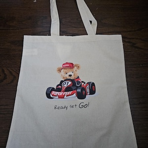 May include: Beige tote bag featuring a teddy bear in a red racing kart. The bear wears a red cap and the kart has the words "Super Fast" and "Ready set Go!" printed on it. The bag has two handles.