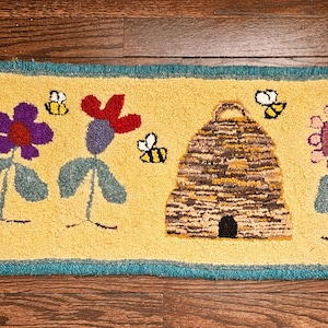Puede incluir: Una alfombra amarilla y azul con una colmena, flores y abejas. Las flores son moradas, rojas y rosas. Las abejas son amarillas y negras.