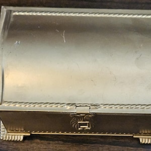 May include: A gold-colored, rectangular jewelry box with a curved lid. The box features a rope-like trim around the lid and base, with decorative feet. The front has a clasp with an ornate design.