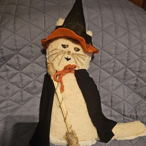 May include: A plush cream-colored cat doll dressed as a witch, wearing a black and orange pointed hat and a black cape. The cat features embroidered facial details, including eyes and whiskers, and holds a small broomstick made of twigs and twine.