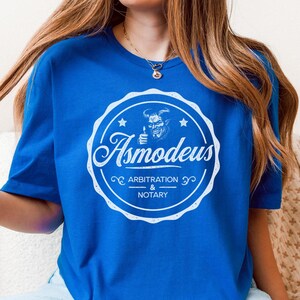 May include: A royal blue t-shirt with a white distressed circle graphic featuring a cartoon devil with horns and a lit candle. The text "Asmodeus Arbitration & Notary" is inside the circle.