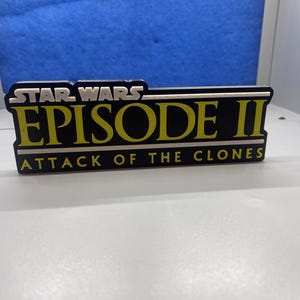 May include: A black and yellow sign with the text "Star Wars Episode II Attack of the Clones".