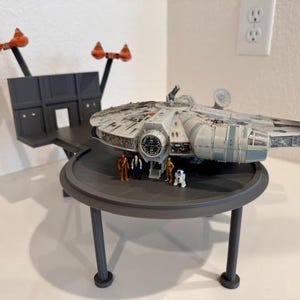 May include: A detailed model of the Millennium Falcon spaceship, with miniature figures of characters, displayed on a dark gray platform with a matching stand. The model includes orange and black accents.