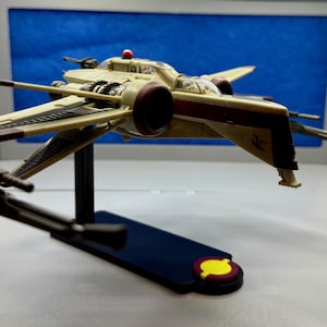 May include: A model starfighter in tan and maroon, mounted on a black stand with a blue base. The starfighter has detailed wings and a cockpit. The base has a yellow and red emblem. The background is blue.