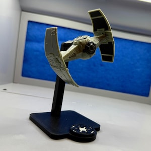 May include: A model of a TIE fighter spacecraft, a vehicle from the Star Wars franchise. The spacecraft is light gray with black solar panels and is mounted on a black stand with a circular base featuring a white emblem.