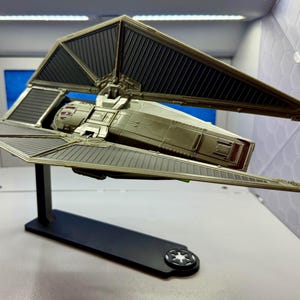 May include: A model of a TIE fighter spacecraft, featuring a central cockpit section and two solar panel wings. The spacecraft is primarily metallic silver and black, mounted on a black stand with a circular emblem. The model is a collectible item.