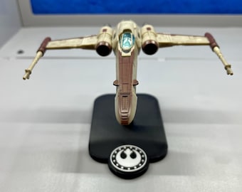 Puede incluir: Maqueta de caza estelar con detalles en beige y marrón, alas extendidas. Montada sobre una base negra con un emblema circular. La cabina es visible con un interior azul-verdoso. Objeto de colección.