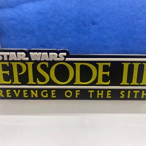Op de afbeelding: Een zwart en geel Star Wars Episode III: Revenge of the Sith logo op een witte achtergrond.