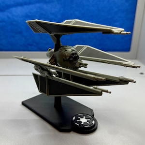 Könnte beinhalten: Ein Miniaturmodell eines TIE-Jägers, eines fiktiven Raumschiffs aus einem Science-Fiction-Franchise. Das Modell ist hauptsächlich grau und schwarz, mit Solarpanelen, die sich vom zentralen kugelförmigen Cockpit erstrecken. Es ist auf einem schwarzen Sockel mit einem kreisförmigen Emblem montiert.