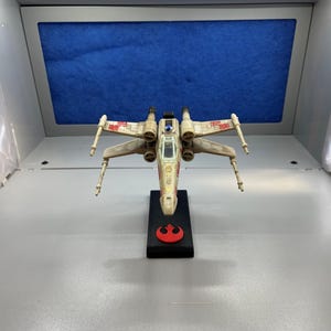 May include: A white and red X-Wing Starfighter model on a black stand with the Rebel Alliance symbol. The model is detailed with a cockpit and wings.