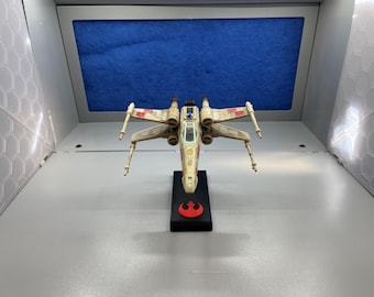 Puede incluir: Un modelo de caza estelar X-Wing Starfighter blanco y rojo sobre una base negra con el símbolo de la Alianza Rebelde. El modelo está detallado con una cabina y alas.