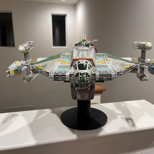 Könnte beinhalten: Ein detailliertes Modell eines grün-weißen Raumschiffs mit orangefarbenen Akzenten. Das Raumschiff steht auf einem schwarzen Ständer und hat ein Cockpit mit einer klaren Kanzel.