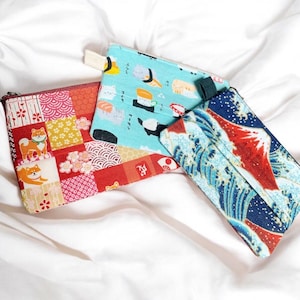 以下が含まれることがあります： 3つの小さな長方形の布製ポーチ。1つは柴犬のパッチワークデザイン、もう1つは寿司柄、3つ目は波と山の風景が描かれています。ポーチは赤、青、白の色合いです。