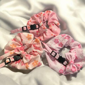 Könnte beinhalten: Drei rosa Stoff-Haargummis mit Blumenmustern und schwarzen verstellbaren Bändern. Jedes Haargummi hat eine kleine schwarze Plastik-Schnalle mit einem Katzenkopf-Design.