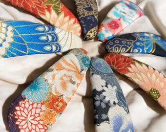 May include: A collection of eight fabric-covered hair clips, each with a unique floral or patterned design. The clips feature a variety of colors, including red, blue, orange, and gold, and are arranged in a circular pattern on a white surface.