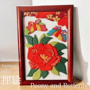 以下が含まれることがあります： 押絵の技法を用いた、赤い牡丹の花と2匹のカラフルな蝶が描かれた手作りのお飾りです。ダークウッドのフレームに収められており、画像の下には「押絵」と「Peony and Butterfly」の文字が書かれています。
