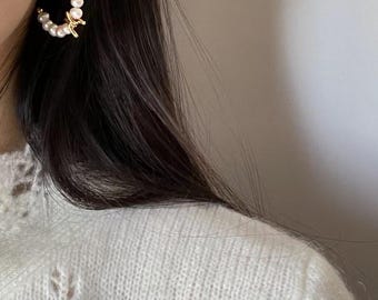 May include: A close-up of a pearl hoop earring with a gold accent. The earring is adorned with small, round pearls. The earring is on a person's ear, with dark hair and a white sweater in the background.