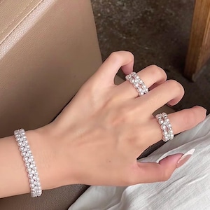 Puede incluir: Una pulsera y dos anillos con pequeñas perlas blancas redondas. La pulsera está en una muñeca y los anillos en los dedos. La joyería se presenta sobre un fondo neutro.