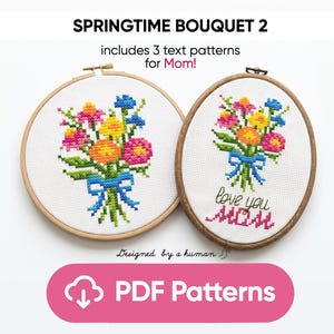 Könnte beinhalten: Zwei Kreuzstich-Stickrahmen mit Blumensträußen. Einer ist rund, der andere oval, beide mit Holzrahmen. Die Sträuße enthalten rosa, orange, gelbe und blaue Blumen, die mit einem blauen Band gebunden sind. Der ovale Reifen sagt "love you MOM".