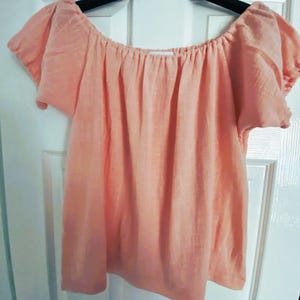 May include: A peach-colored off-the-shoulder top with short, puffed sleeves. The top is made of a soft, lightweight fabric.
