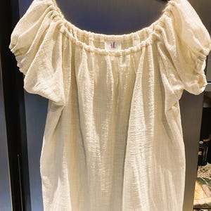 May include: Off-white, short-sleeved top with a gathered neckline and puffed sleeves. The fabric has a textured appearance. The top is hanging on a black hanger against a neutral background. A small tag is visible near the neckline.