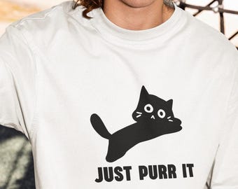 May include: White t-shirt with a black cat graphic and the text "JUST PURR IT". The shirt is paired with yellow shorts and blue sunglasses. The person is sitting on a playground structure.