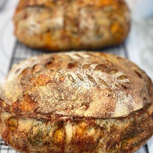 May include: Two loaves of freshly baked sourdough bread with a golden brown crust and a slightly cracked surface. The bread has a rustic appearance and is likely made with a sourdough starter.