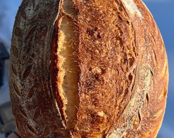 May include: A close-up of a freshly baked loaf of sourdough bread with a golden brown crust and a decorative leaf pattern. The bread has a deep crack down the center, revealing a soft, airy interior.