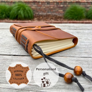 Könnte beinhalten: Ein braunes Lederjournal mit Wickelverschluss und schwarzem Band-Lesezeichen. Das Journal ist mit "Genuine 100% Leather" und "Personalized gift" beschriftet. Das Journal liegt auf einer Holzoberfläche.