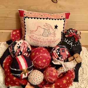 May include: A decorative fabric display featuring a rectangular pillow with a rabbit illustration and text, surrounded by several small, round, fabric-covered spheres in red, white, and blue patterns, a black star ornament labeled USA, and frayed fabric accents.