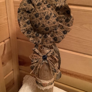 Könnte beinhalten: Eine dekorative Glasflasche, gekrönt von einer großen, runden Stoffblume mit beigem und schwarzem Blumenmuster. Der Flaschenhals ist mit einer Juteschnurschleife, einem dunkelblauen Knopf und einem Stoffstreifen mit kleinem Holzherz verziert.
