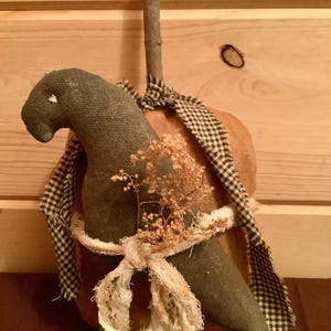 May include: A fabric pumpkin decoration featuring a dark green cloth bird figure attached to the front. The pumpkin has a brown twig stem and is accented with a black and white checkered fabric strip and a frayed cream-coloured ribbon tied around it.