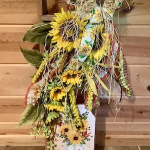 May include: A decorative wall hanging featuring a wooden sign with a mason jar and sunflower illustration, printed with the text "HELLO fall". The sign is adorned with a large arrangement of artificial sunflowers, green leaves, and ribbons.