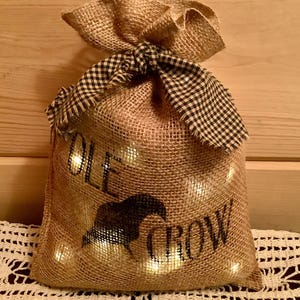 May include: A tan burlap drawstring bag featuring a black silhouette of a crow and the text OLE CROW printed on the front. The bag is tied at the top with a black and white checkered fabric ribbon and contains internal warm white lights.
