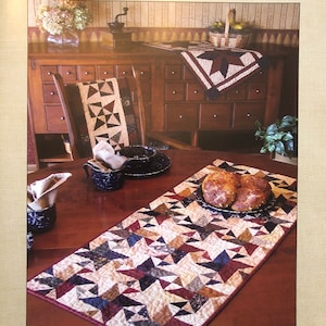 May include: A table runner with a geometric pattern in shades of red, blue, brown, and white. The runner is made of fabric and has a quilted texture. The runner is laid out on a wooden table with a bowl of bread rolls and a napkin. The text "Mostly Table Runners Three" and "Calico Printworks" is visible in the image.