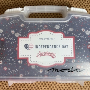 May include: A translucent plastic storage case with a white handle, featuring a dark blue floral pattern and a central label that reads "moda INDEPENDENCE DAY Sweetwater" with a heart-shaped American flag icon and the word "moda" below.