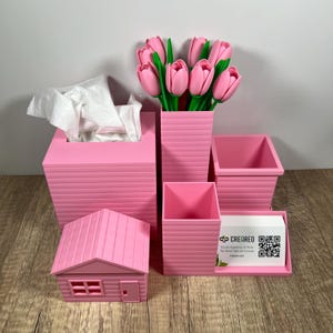 May include: A pink desk organizer set includes a tissue box, a vase with pink tulips, a small house, and several containers. A business card holder with a QR code is also included. The set is made of a ridged material.